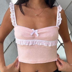 May include: A pink crop top with white lace trim and a white bow at the center. The top has a ribbed texture and a square neckline.