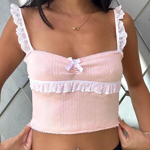 May include: A pink crop top with white lace trim and a white bow at the center. The top has a sweetheart neckline and thin straps.