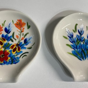 May include: Two white ceramic spoon rests with painted floral designs. One spoon rest features blue and white flowers, while the other features blue, yellow, red, and orange flowers.