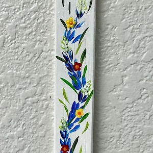 May include: A white wooden bookmark with a hand-painted floral design featuring blue, yellow, and red flowers with green leaves. The bookmark has a hole at the top with a blue ribbon attached, allowing it to hang from a hook on a textured wall.