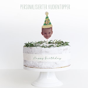 Puede incluir: Un pastel blanco decorado con ramitas de romero verde y un adorno de pastel personalizado. El adorno presenta la cara de un ni&ntilde;o con un gorro de fiesta verde y amarillo con el n&uacute;mero 6. El pastel est&aacute; decorado con las palabras "Feliz cumplea&ntilde;os".