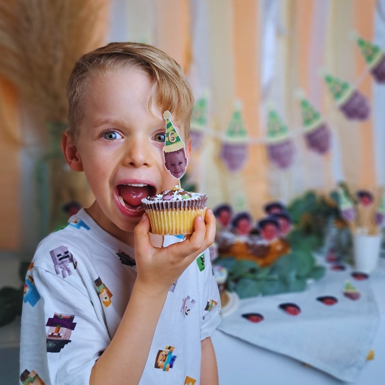 Puede incluir: Una persona joven que lleva una camisa blanca con un dise&ntilde;o de Minecraft est&aacute; sosteniendo un cupcake con una peque&ntilde;a figura en la parte superior. El cupcake est&aacute; decorado con chispas de colores. El fondo est&aacute; decorado con una pancarta de peque&ntilde;as figuras que llevan sombreros de fiesta.