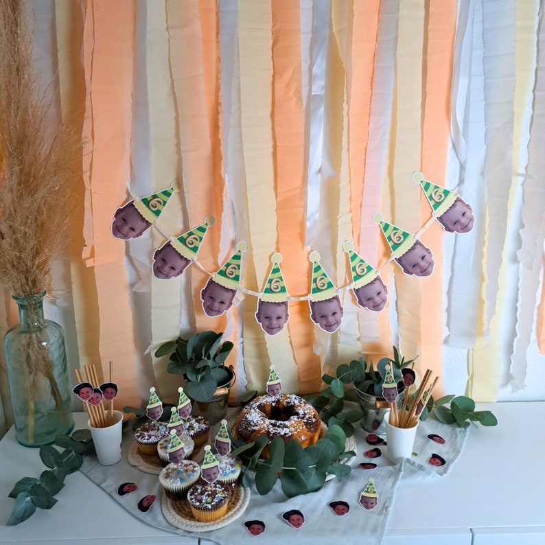 Puede incluir: Una decoraci&oacute;n de fiesta de cumplea&ntilde;os con una pancarta de fotos de ni&ntilde;os con gorros de fiesta. La pancarta est&aacute; colgada en una pared decorada con serpentinas de papel de seda de color melocot&oacute;n y crema. Hay cupcakes con glaseado y chispas en una mesa delante de la pancarta.