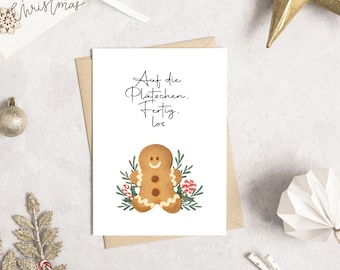 May include: A white greeting card with the text "Auf die Plätzchen. Fertig. Los" and a gingerbread man illustration. The card is on a neutral surface with Christmas decorations, including a gold star and a "Merry Christmas" tag.