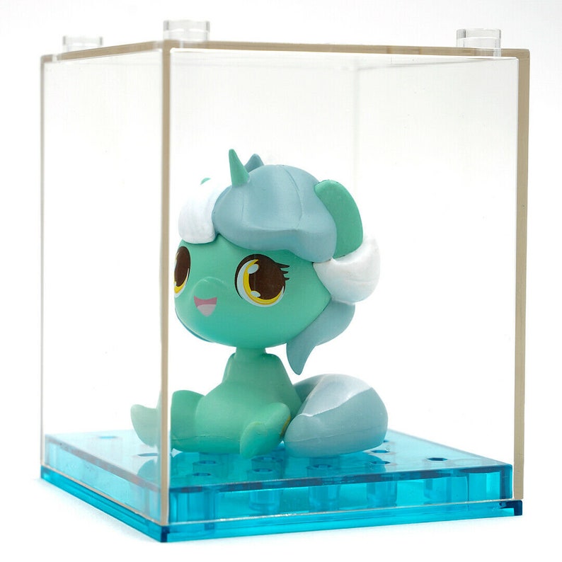 May include: A teal figurine with white and light blue accents, displayed in a clear acrylic case with a blue base. The figurine has large brown eyes with yellow highlights and a smiling expression.
