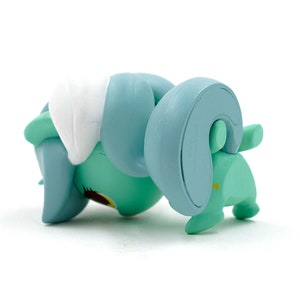 May include: A turquoise figurine of a stylized animal, possibly a ram or sheep, with large curved horns and a white patch on its back. The figurine is lying on its side, showcasing its rounded form and simple design.