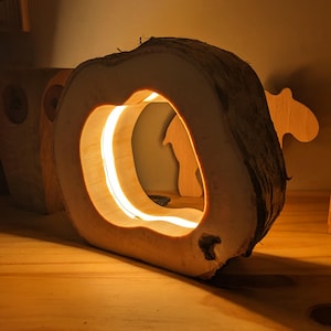 Puede incluir: Lámpara de madera hecha a mano con un brillo cálido. La lámpara presenta una sección grande y ahuecada, creando una abertura central. La madera tiene un aspecto natural y texturizado, y la fuente de luz es visible dentro de la abertura.