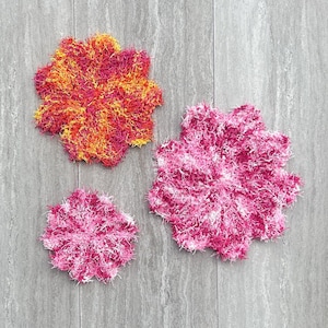 May include: Three handmade, flower-shaped crochet items in varying sizes. The largest is a mix of orange, yellow, and pink. The other two are shades of pink and white. These decorative items are likely for home decor.