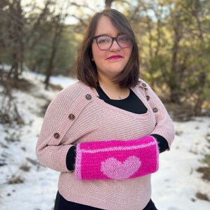 Puede incluir: Una mujer con un suéter rosa con botones en la manga y un calientamanos de punto rosa en forma de corazón. Lleva gafas y tiene el pelo castaño.