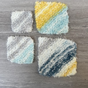 May include: Four handmade, square washcloths in varying sizes and color patterns. The washcloths feature a textured, looped design with diagonal stripes in shades of gray, white, yellow, and blue. The washcloths are arranged on a gray surface.