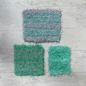 May include: Three handmade crochet scrubbies in shades of green and gray. The largest is rectangular with horizontal stripes of teal and gray. The other two are square, one in solid green and the other in a mix of green and gray.