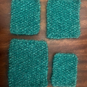 May include: Four teal-coloured, square-shaped scrubbies or washcloths. The items have a textured, looped appearance, and are arranged on a dark wooden surface. The scrubbies vary slightly in size, with one being noticeably smaller.
