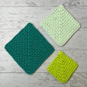 May include: Three handmade, square crochet washcloths in shades of green and yellow. The largest is a deep teal, the next is a light green, and the smallest is a bright yellow-green. The washcloths are arranged on a light grey and white wood-grain surface.