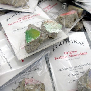 May include: A pile of small, gray concrete pieces, each encased in a clear plastic bag with a white card. The cards have the text "Original Berliner Mauer-Stein" and "Ein Stück deutscher Geschichte" printed on them. The text on the cards is in German.