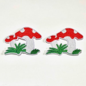 May include: Two identical embroidered patches featuring a pair of mushrooms with red caps, white spots, and white stems, set against a base of green grass. The design is rendered in vibrant thread colours with a distinct white border outlining the shapes.