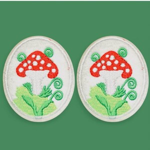 May include: Two circular embroidered patches featuring a red mushroom with white spots on a white background. The mushroom is surrounded by green embroidered leaves and swirling vine details. Each patch has a white stitched border edge.