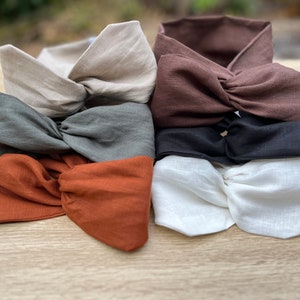 May include: A stack of six fabric headbands in various colours, including beige, brown, grey, orange, black, and white. The headbands are made of a soft, textured fabric and are tied in a knot at the top.