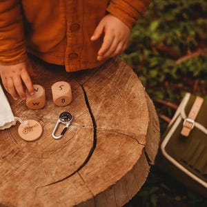 Puede incluir: Un conjunto de dos bloques de madera con letras, una ficha de madera circular con el texto Compass Set, una pequeña brújula de mosquetón plateada y una bolsa de tela blanca con cordón, dispuestos sobre un tocón de árbol.