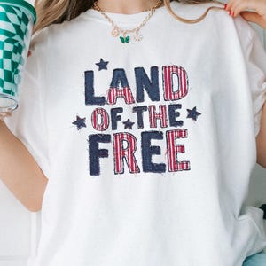 May include: A white short-sleeved t-shirt featuring the text LAND OF THE FREE in a distressed, red and blue plaid pattern. The design includes small navy blue stars interspersed between the words. The shirt is made of a plain white fabric.