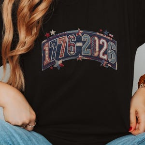 May include: A black short-sleeved t-shirt featuring a graphic print of the text 1776-2026 in a distressed, multi-colored pattern with small star accents. The design is arched across the chest, incorporating red, white, and blue tones.