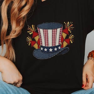 May include: A black short-sleeve t-shirt featuring a graphic of a top hat with red and white stripes, a blue brim with white stars, and yellow fireworks on each side. The design has a textured, fabric-like appearance on the black cotton fabric.