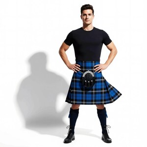 A man wearing a black t-shirt and a blue and black plaid kilt stands with his arms crossed, with a serious expression on his face.