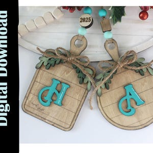May include: Two wooden ornaments, one square and one round, with teal letters and a wood grain pattern. The square ornament has the letters "CH", and the round ornament has the letter "A". Both are decorated with twine and faux greenery. A beaded garland with the year "2025" is visible.