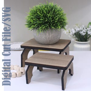 May include: Two wooden plant stands with a square top and curved legs. The stands are different sizes, with a small green plant in a gray pot on the larger stand. The text "Digital Cut File/SVG" is on the left side.