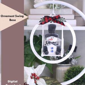 May include: A white metal ornament swing base with a snowman ornament. The snowman has a black top hat, a black and white checkered scarf, and blue button accents. A red and black plaid bow and greenery adorn the top of the swing. The text "Ornament Swing Base" and "Digital Download" are visible.