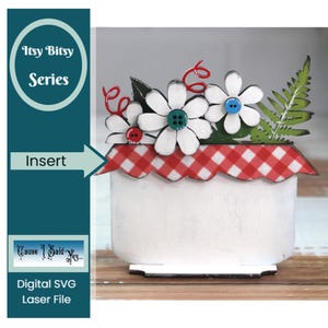 May include: A white ceramic flower pot with a red and white checkered rim. The pot is decorated with a bouquet of white flowers with red and blue buttons in the center of each flower. A green fern is on the right side of the pot.