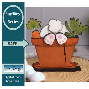 May include: A wooden cutout of a white bunny rabbit sitting in a brown flower pot with green leaves and an orange carrot. The bunny's back feet are visible and the pot is sitting on a black base.