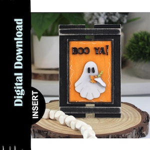 May include: A wooden Halloween decoration featuring a white ghost holding a small orange treat, set against an orange background with the words "BOO YA!". The frame is black, and the decoration sits on a wooden base.