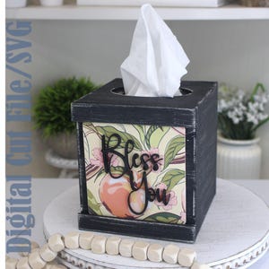 May include: A square black wooden tissue box cover with a distressed finish. The front panel features a botanical print with peaches and pink flowers, overlaid with black script lettering that reads Bless You. A white tissue protrudes from the top.