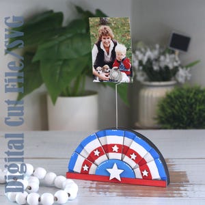 May include: A photo holder with a red, white, and blue design. The holder has a semi-circular base with star accents and a wire to hold a photo. The image shows a photo of a woman and child.