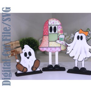 May include: Three ghost figures on black stands. The left ghost holds a sign reading "Trick or Treat" and "Smell My Feet". The center ghost has a colorful patchwork pattern and holds a cup. The right ghost is white with an orange bow.