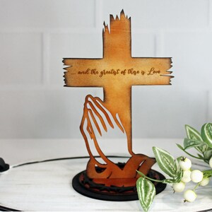 Puede incluir: Cruz de madera con manos rezando recortadas, con el texto "...and the greatest of these is Love". La cruz es de color marr&oacute;n c&aacute;lido, con un dise&ntilde;o de borde desgastado. La base es un c&iacute;rculo marr&oacute;n oscuro.