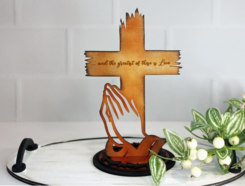 Puede incluir: Cruz de madera con manos rezando recortadas, con el texto "...and the greatest of these is Love". La cruz es de color marr&oacute;n c&aacute;lido, con un dise&ntilde;o de borde desgastado. La base es un c&iacute;rculo marr&oacute;n oscuro.