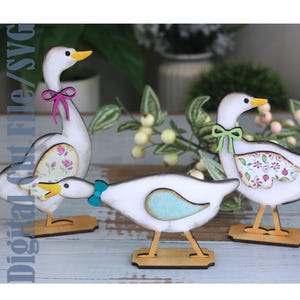 May include: Three wooden goose figurines with white bodies, yellow beaks and feet, and decorative bows. The geese have floral patterned wings and stand on wooden bases. The background includes greenery and a blurred vase.