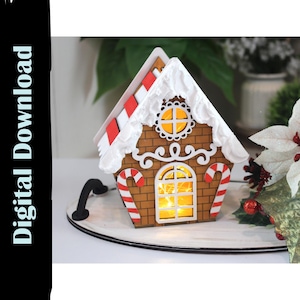 Puede incluir: Una casa de jengibre decorativa con interior iluminado, diseño de ladrillos marrones, glaseado blanco y detalles de bastones de caramelo rojos y blancos. La casa está sobre una bandeja blanca, con el texto "Digital Download".