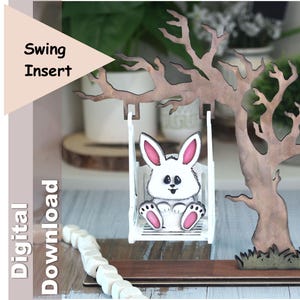 Puede incluir: Un inserto de columpio de madera con un conejito blanco con detalles rosas, sentado en un columpio blanco. El columpio está suspendido de un árbol estilizado. Se ven las palabras "Swing Insert" y "Digital Download".