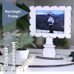 May include: A white rectangle frame displaying a photograph of skiers in a mountain landscape. The frame sits on a white stand with a decorative base. A small potted plant and the text "Digital Download" are also visible.