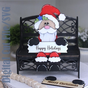May include: A layered wooden cutout of a Santa Claus figure wearing a red hat and white beard, sitting on a black metal bench. The figure holds a white sign that reads Happy Holidays. A separate wooden sign reads Merry Christmas.