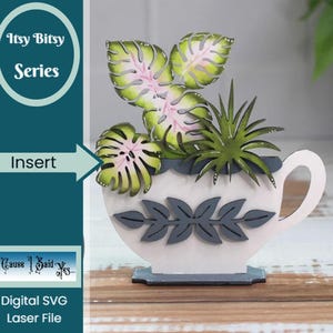 May include: A white ceramic teacup with a handle, decorated with gray leaves, is filled with green and pink leafy plants. The teacup is sitting on a wooden surface. The text "Itsy Bitsy Series" is in a circle above the teacup and the text "Insert" is in a rectangle to the left of the teacup. The text "Cause I Said Yes..." is in a rectangle below the teacup and the text "Digital SVG Laser File" is below that.