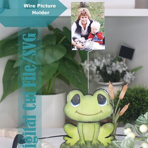 May include: A whimsical wire picture holder featuring a wooden frog figurine. The frog is painted green with black outlines and holds a photo of a woman and child. The text "Digital Cut File/SVG" is visible.