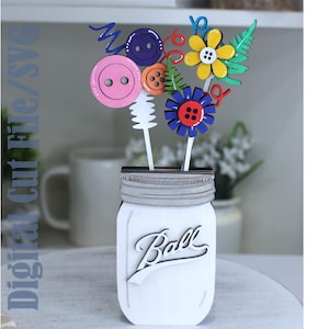 May include: A white wooden mason jar cutout with the word Ball in script, containing a bouquet of colorful button flowers in pink, purple, orange, yellow, and blue, accented with green fern leaves and red swirls. Digital Cut File/SVG text is visible.