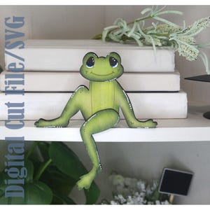Puede incluir: Una figura de rana verde con una cara sonriente y detalles blancos. La rana está posada en un estante blanco, con el texto "Digital Cut File/SVG" visible a la izquierda. Los brazos y las piernas de la rana están extendidos.