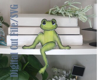 Peut inclure: Une figurine de grenouille verte avec un visage souriant et des détails blancs. La grenouille est posée sur une étagère blanche, avec le texte "Digital Cut File/SVG" visible à gauche. Les bras et les jambes de la grenouille sont tendus.