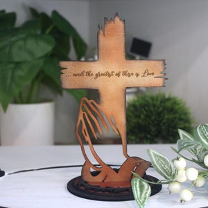 Puede incluir: Cruz de madera con la inscripci&oacute;n "...and the greatest of these is Love". La cruz tiene un aspecto r&uacute;stico y desgastado, con una base estilizada. Un art&iacute;culo decorativo.