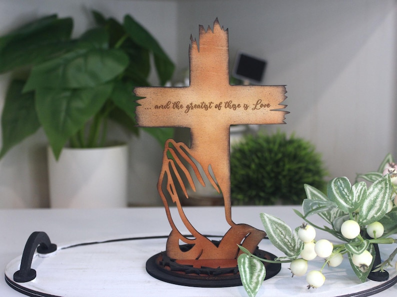 Puede incluir: Cruz de madera con la inscripci&oacute;n "...and the greatest of these is Love". La cruz tiene un aspecto r&uacute;stico y desgastado, con una base estilizada. Un art&iacute;culo decorativo.