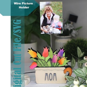 May include: A wire picture holder with a photo of a woman and child with puppies. The holder features a wooden flower pot with colorful tulips and the word "MOM". The text "Digital Cut File/SVG" is also present.